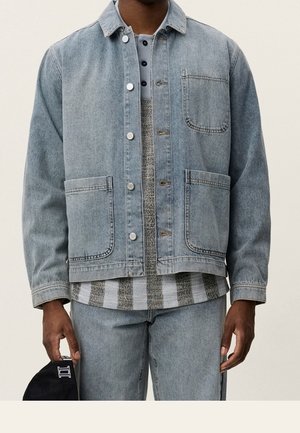 Mężczyzna w jasnej kurtce dżinsowej i dżinsach, trzymający czarną czapkę, nałożoną na pasiastą szaro-białą koszulę.