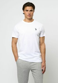 Vit bomullströja med rund hals, korta ärmar och ett litet svart emblem på vänster bröst. Kombinerad med gråa mjukisbyxor.