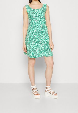 Robe à imprimé floral vert avec un décolleté arrondi, sans manches, et un ourlet évasé. Associée à des sandales à plateforme blanches.