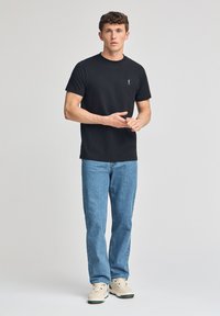 T-shirt nera in cotone a maniche corte con un piccolo logo sul petto, abbinata a jeans dritti blu chiaro e sneakers beige.