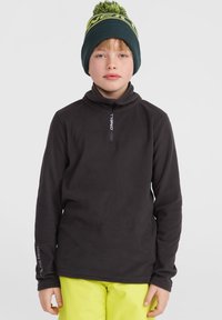 Pull en polaire noir avec un col zippé mi-haut et le logo Polartec sur la manche. porté avec un pantalon jaune vif et un bonnet vert avec un pompon.