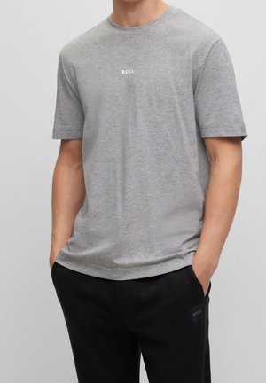 Mand iført en ensfarvet grå kortærmet t-shirt med "BOSS" logo og sorte bukser med hænderne i lommerne mod en ensfarvet baggrund.