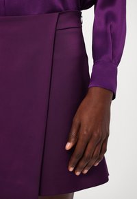 Main à la peau foncée reposant à côté d'une jupe violette et d'une chemise à manches longues assortie avec des poignets, sur un fond blanc.