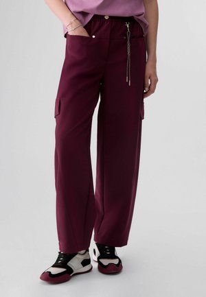 Persona che indossa pantaloni cargo bordeaux larghi, sneakers bianche e nere e una maglietta rosa con una mano in tasca su uno sfondo semplice.