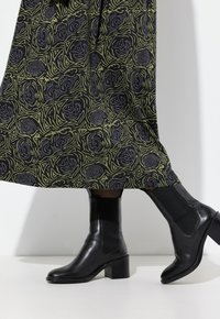 Černé kožené kotníkové boty s robustním podpatkem a elastickými bočními panely, obuté s zeleno-černou sukni s vzory a vířícími designy.