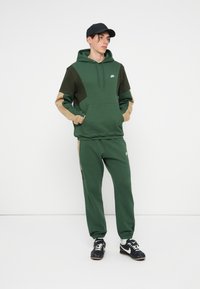 Vihreä huppari, jossa on erottuvat tummanvihreät ja vaaleanruskeat paneelit, edessä tasku, yhdistettynä samanvärisiin vihreisiin collegehousuihin ja mustiin urheiluvarusteisiin.