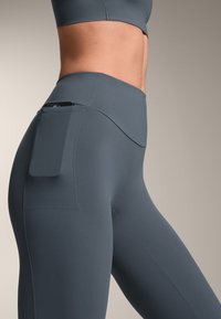 Hoch geschnittene, figurbetonte Leggings aus dunkelgrauem Stoff, ausgestattet mit einer Seitentasche und einer glatten Textur, die minimale Nähte und klare Linien aufweist.