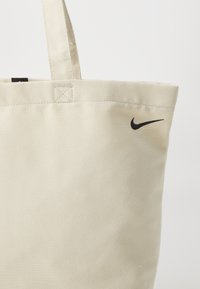 Borsa a spalla in tela beige con logo Nike nero. Presenta cuciture robuste e un design semplice. Due manici per un facile trasporto.
