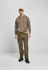 Felpa marrone con tasca, indossata sopra una maglietta gialla; pantaloni cargo oliva con tasche laterali; cappellino beige; scarpe nere con suole texture.