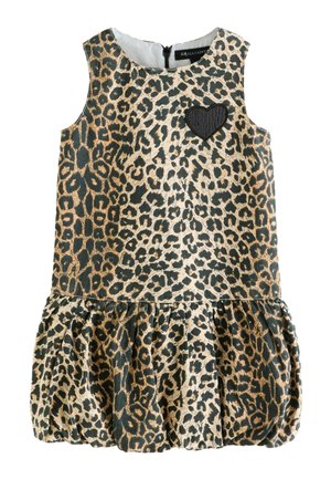 Ragazza giovane in piedi, indossa un vestito smanicato con stampa leopardata e una toppa a forma di cuore, e sandali neri con fibbie, su uno sfondo chiaro e uniforme.