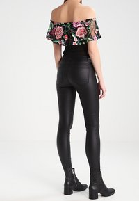 Femme portant un pantalon noir en simili cuir, des bottines noires, et un haut décolleté avec une broderie florale colorée, de dos.