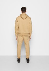 Sudadera beige con capucha de corte holgado y puños acanalados, combinada con pantalones ajustados a juego. El material parece suave con una textura lisa.