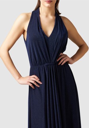 Femme portant une robe bleu marine sans manches à col halter en V avec taille cintrée, mains posées sur les hanches devant un fond neutre.