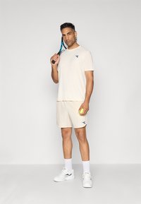 Light beige athletic t-shirt and shorts, featuring a blue logo. The figure holds a tennis racket and a neon yellow tennis ball. White sneakers.