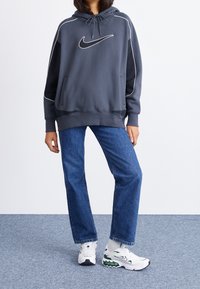 Gray Nike hoodie with white accents, kangaroo pocket, paired with blue straight-leg jeans and white-green sneakers on a textured floor.