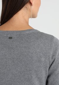 Grå strikket sweater med rund hals, ribbede manchetter og et lille logo mærke på bagsiden. Glat tekstur og tætsiddende design.