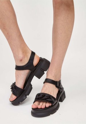 Czarne sandały na grubym obcasie z łańcuchem i paskami na kostkę noszone na bare stopach na tle jednolitego białego tła.