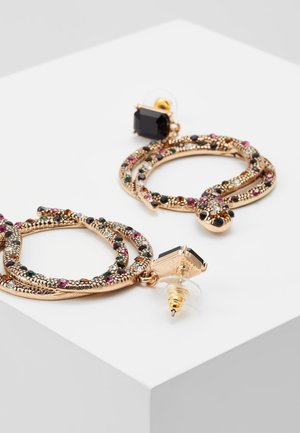 Boucles d'oreilles créoles en forme de serpent doré avec pierres multicolores et clous carrés noirs sur une surface blanche.