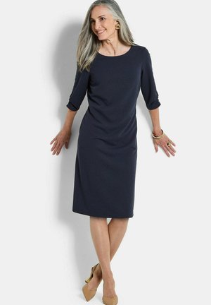 Frau mit grauen Haaren, die ein marineblaues knielanges Kleid, beige Absätze, goldene Ohrringe, Armband und Ring trägt, lächelnd an eine weiße Wand gelehnt.