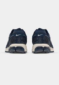 Nike sportbačiai tamsiai mėlynos spalvos su tekstūruotu tinkliniu paviršiumi, sidabriniais akcentais ir kreminės spalvos padu. Yra raštuota kulno dalis.