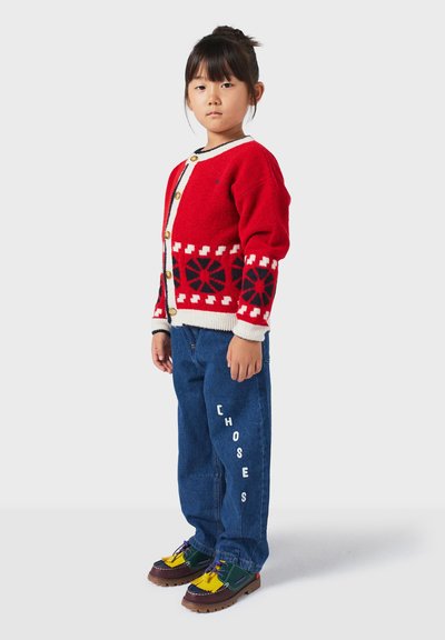 Czerwony sweter robiony na drutach z białymi i granatowymi wzorami, złote guziki, w połączeniu z niebieskimi dżinsami z napisem "CHOSES" na biało oraz wielokolorowymi butami.