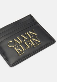 Černý kožený držák karet s několika sloty, vyznačující se výrazným zlaceným embosovaným logem "CALVIN KLEIN" a textem "NEW YORK CITY" na spodní straně.