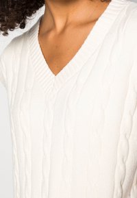 Weißer Strickpullover mit V-Ausschnitt, kurzen Ärmeln und einem strukturierten Zopfmuster. Weiches Material mit gerippten Details am Ausschnitt.
