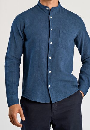 Homme portant une chemise bleu foncé à manches longues avec col mandarin et poche poitrine, associée à un pantalon foncé.