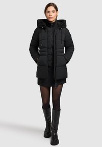 Jachetă puffer neagră cu glugă, închidere cu fermoar și șnururi reglabile. Prezintă o textură matlasată și buzunare laterale, asociată cu o fustă neagră și cizme.