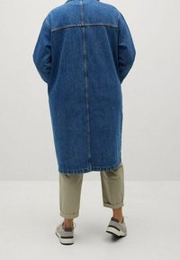 Person som bär en lång blå denimjacka, beige uppvikta byxor och vita sneakers, står med händerna i fickorna och vänder ryggen mot oss.