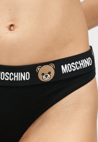 Κοντινό πλάνο στη μέση ενός ατόμου που φορά μαύρα εσώρουχα με λωρίδα μέσης με λογότυπο που εμφανίζει πρόσωπο αρκούδας και τη λέξη "MOSCHINO" επαναλαμβανόμενα.