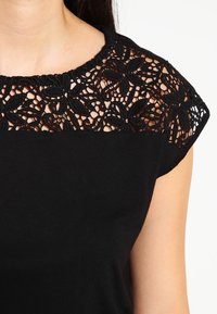 Czarny top z koronkową nakładką w okolicy dekoltu z kwiatowym wzorem. Krótkie rękawy i gładka faktura tkaniny na całej powierzchni.