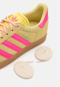 Baskets en daim jaune avec accents roses, dotées de trois bandes roses, semelle texturée et deux paires de lacets plats en blanc et rose. Modèle : Gazelle.