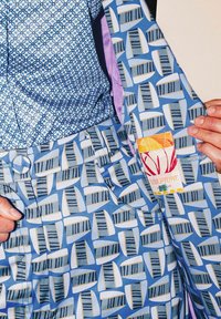 Shorts à motifs bleus avec des rayures verticales et des dessins de voiles, comportant une étiquette en tissu portant le nom "Sculpture" avec des accents orange et jaune.