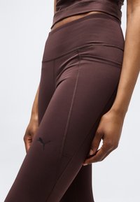 Κοντινό πλάνο ενός ατόμου που φοράει σκουρόχρωμα, ψηλόμεσα κολάν Puma σε καφέ σκούρο χρώμα, με λεπτομερή ραφές και λογότυπο στο πλάι του μηρού.