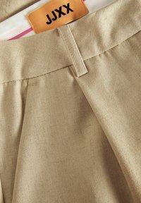 Pantalones plisados beige con una textura suave, que presentan una cinturilla plana y una etiqueta de marca en naranja y negro en el interior.