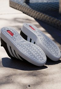 Des baskets noires à semelle blanche avec des accents rouges sont placées semelle vers le haut sur une surface en béton ensoleillée près d’un pavé texturé.