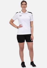 Polo shirt bianco con accenti neri, logo sul lato sinistro, abbinato a pantaloni corti neri. Modello che indossa scarpe da ginnastica nere, in piedi davanti a uno sfondo neutro.