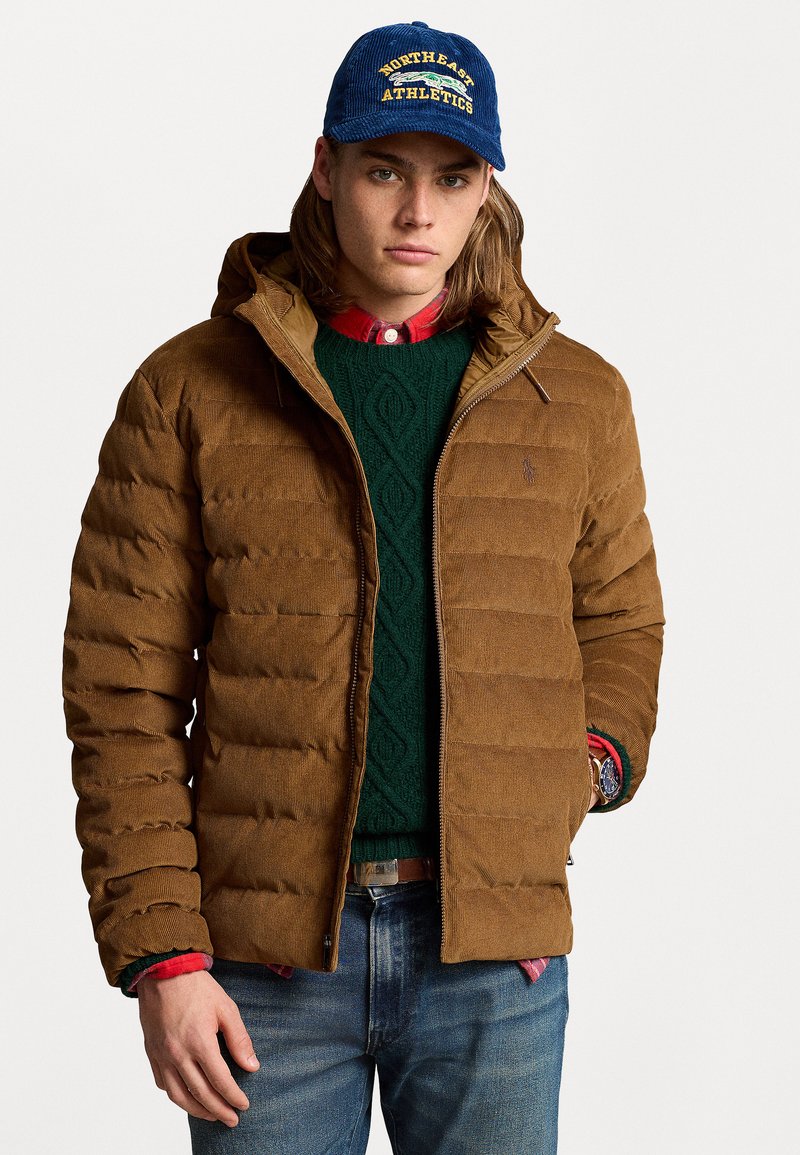 Young man wearing a blue "Northeast Athletics" cap, brown quilted jacket, green cable-knit sweater, red plaid shirt, and blue jeans.