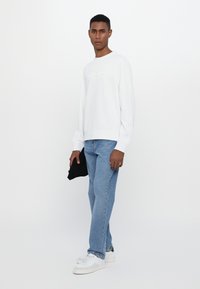 White sweatshirt with embossed text, styled with light blue jeans and white sneakers. Model holds a black item in hand, against a white background.