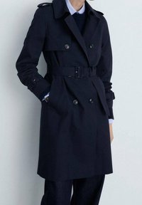 Osoba oblečená v tmavě modrém trench coat s páskem a dvouřadými knoflíky, stojící s jednou rukou v kapse na světlém pozadí.