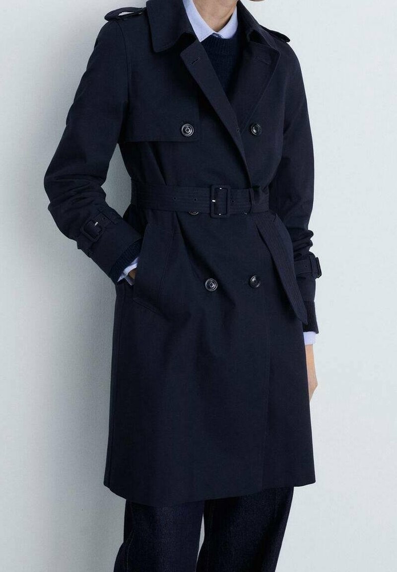 Osoba oblečená v tmavě modrém trench coat s páskem a dvouřadými knoflíky, stojící s jednou rukou v kapse na světlém pozadí.