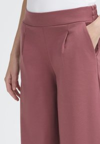 Närbild av en person som har på sig högmidjade, dammrosafärgade vida byxor med sidofickor, där en hand är i fickan, mot en enfärgad vit bakgrund.