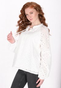 Weiße bestickte Bluse mit Rundhalsausschnitt, Kordelzugdetail, Ballonärmeln und Lochmustern. Kombiniert mit dunklen Jeans.