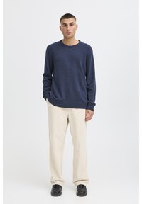 Navy knitted sweater with a crew neck, worn over cream corduroy pants. Sweater features long sleeves and ribbed cuffs, styled casually.