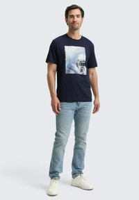 Tricou din bumbac de culoare albastru închis, cu un imprimeu grafic al unui vehicul pe un fundal snowy. Asortat cu blugi de culoare albastru deschis și adidași albi.