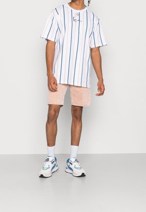 Chemise blanche à manches courtes avec rayures bleues et rouges, associée à un short en velours côtelé rose clair et des baskets de sport multicolores.
