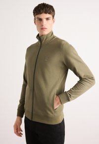Felpa zip-up verde oliva con colletto alto, polsini a coste e vestibilità rilassata. Presenta una piccola toppa con logo sul petto e tessuto testurizzato.