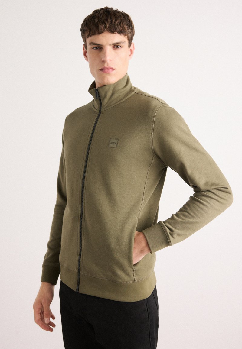 Felpa zip-up verde oliva con colletto alto, polsini a coste e vestibilità rilassata. Presenta una piccola toppa con logo sul petto e tessuto testurizzato.