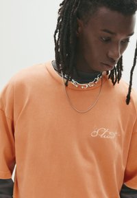 Orange bomulds T-shirt med en afslappet pasform, med et lille hvidt logodesign på brystet. Laget med en sort polo under.